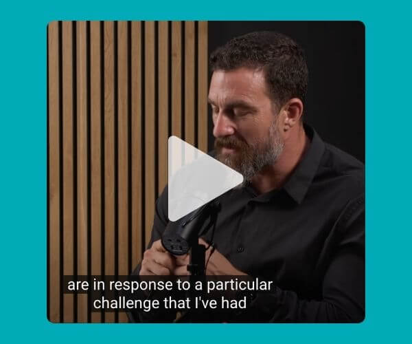 A man with a beard and dark shirt speaks into a microphone in a podcast-style setup. Wooden paneling is behind him, and the caption at the bottom reads, “are in response to a particular challenge that I’ve had.”