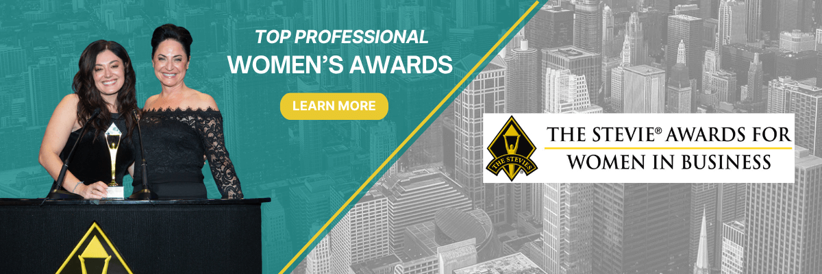 Two women smiling on stage while holding a trophy at a podium, promoting the Stevie Awards for Women in Business with a city skyline in the background and a yellow "Learn More" button.