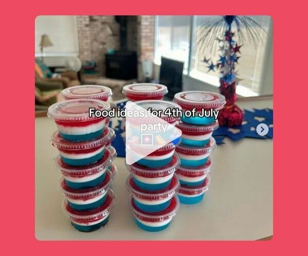 Stacks of red, white, and blue layered desserts in plastic cups sit on a kitchen counter, with text overlaid that says “Food ideas for 4th of July party.” A festive centerpiece with stars is visible in the background, and a play button is centered on the image.