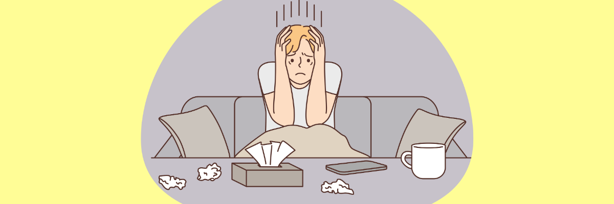 Cartoon illustration of a person sitting on a couch looking stressed, with their head in their hands. There are used tissues, a tissue box, a mug, and a phone on the table in front of them, all set against a pale yellow background.