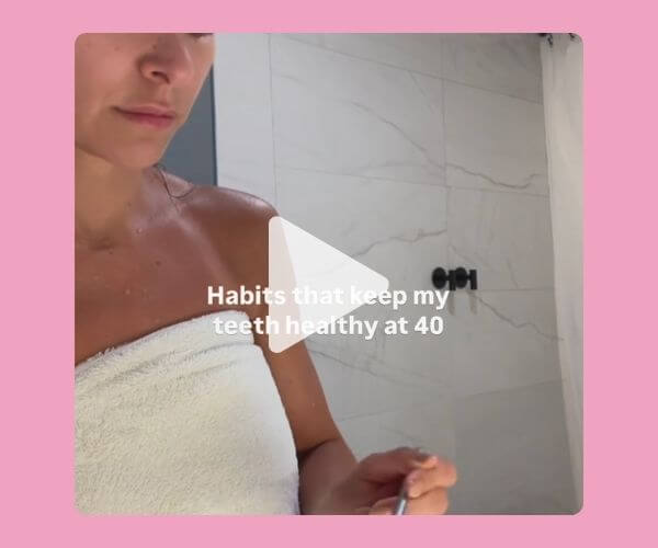 Woman wrapped in a towel in a bathroom with white tile walls, holding a toothbrush with the text overlay: "Habits that keep my teeth healthy at 40.โ