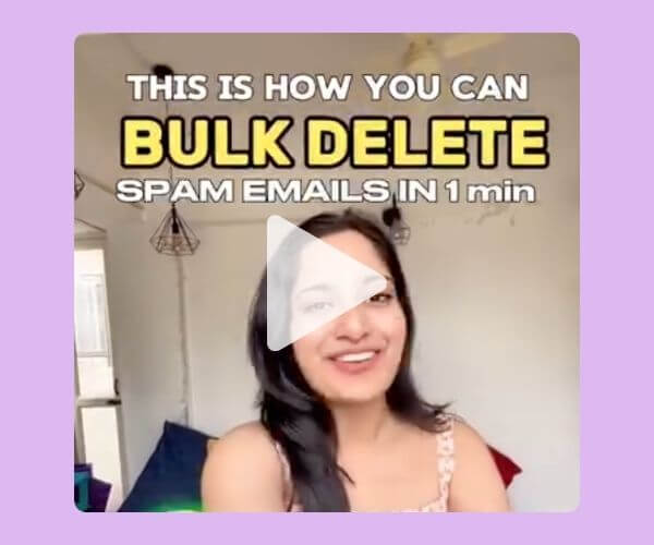 Screenshot of a video featuring a smiling woman with long dark hair, sitting in a bright room with hanging lights. Bold text above her reads, “THIS IS HOW YOU CAN BULK DELETE SPAM EMAILS IN 1 min.” A play button is overlaid on the center of the image.