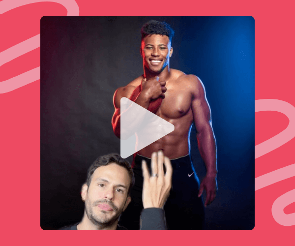 A shirtless man stands confidently against a dark backdrop with red and blue lighting, smiling with one hand resting on his shoulder. In the bottom corner, another man gestures with his hand while looking at the camera.