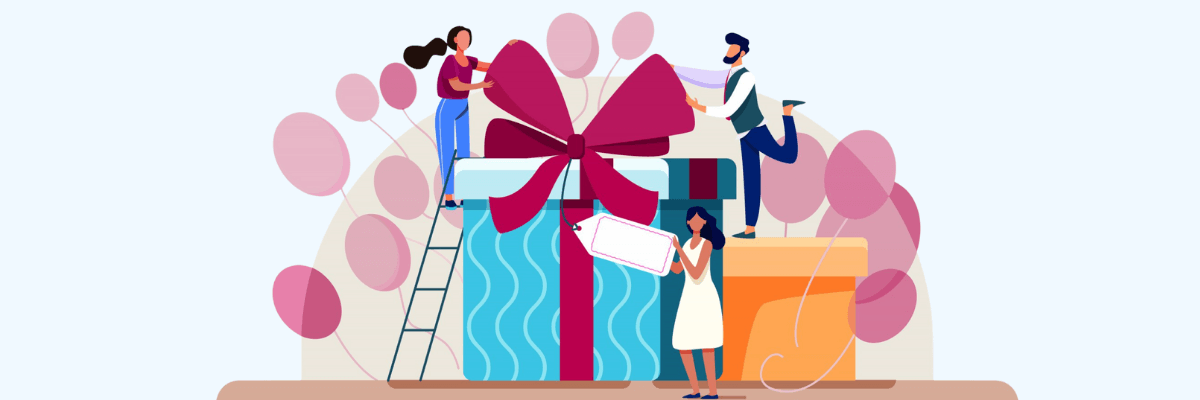 A group of people work together to decorate an oversized gift box—one tying a huge red bow on top, another standing on a ladder, and another holding a blank gift tag—with pink balloons floating all around.