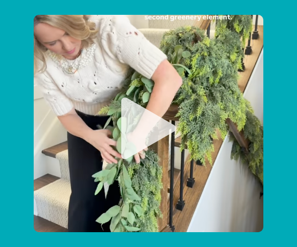 A woman in a cozy sweater decorates a staircase railing with lush greenery, arranging leaves to make a festive garland. It feels very “holiday home makeover” and super crafty.