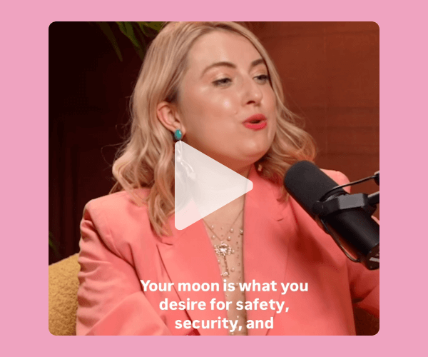 A woman in a coral blazer speaks into a microphone during a podcast recording, set against a warm, softly lit studio background with plants and a pink frame. White on-screen text reads, “Your moon is what you desire for safety, security, and”.