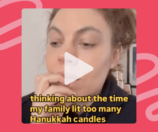 The video frame shows a woman resting her hand against her mouth with the on-screen text reading, “thinking about the time my family lit too many Hanukkah candles.” It’s framed by a bright pink background with playful squiggle patterns.