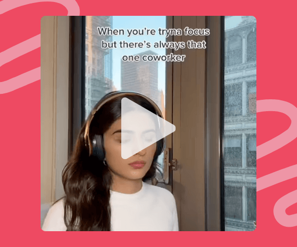 A woman wearing headphones sits by a large window with a city building visible outside against a pink patterned background. Text on the video reads, “When you’re tryna focus but there’s always that one coworker.”