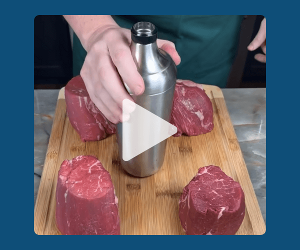 A close-up video still shows a hand holding a stainless steel shaker bottle upright on a wooden cutting board, surrounded by four raw beef steaks arranged around it. The image sits within a dark teal-blue border, with a translucent play button overlay centered on top.