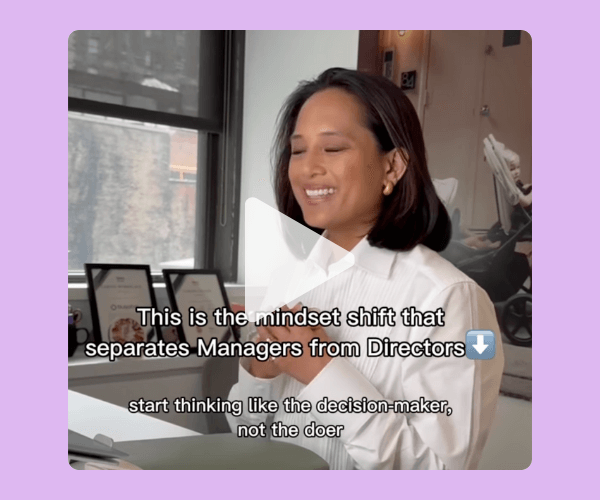 A woman in a white button-down shirt smiles while speaking at a desk in a bright office, with framed photos and a large window showing an urban building behind her. White on-screen text overlays the video reading “This is the mindset shift that separates Managers from Directors,” and the video is framed by a soft lavender background with a play button centered over the image.