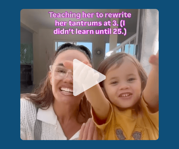 A smiling woman with glasses sits beside a young child in a yellow shirt, both raising their hands toward the camera in a bright living room with stairs in the background. Pink text at the top reads, “Teaching her to rewrite her tantrums at 3. (I didn’t learn until 25.)”.