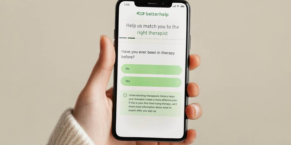 A hand holding a smartphone displaying a BetterHelp sign-up screen that asks questions to match the user with the right therapist. The screen shows green “Yes” and “No” options against a clean, neutral background.