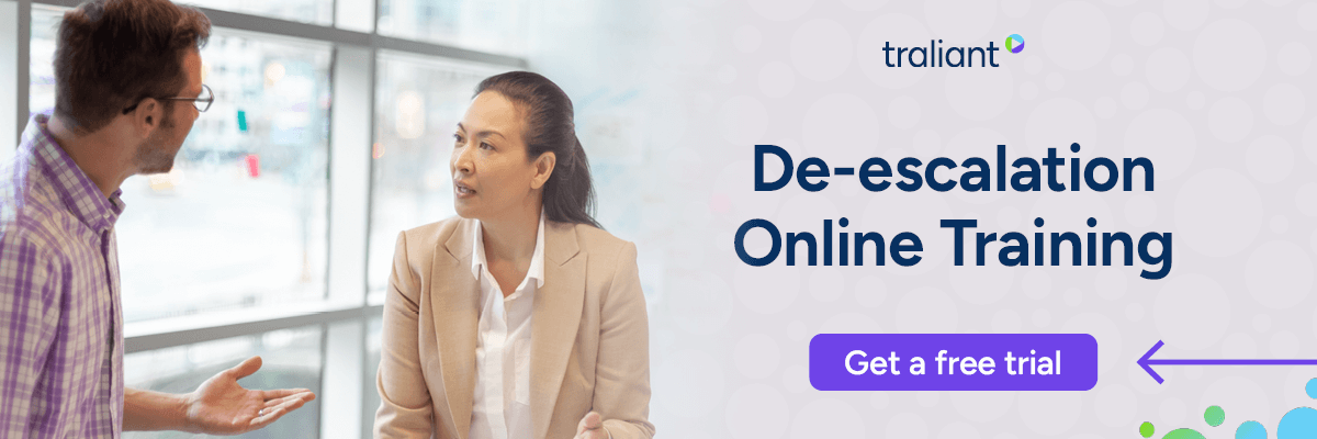 A man gestures while speaking to a woman in a beige blazer near large office windows during a tense conversation. The headline reads “De-escalation Online Training” beside a purple “Get a free trial” button.