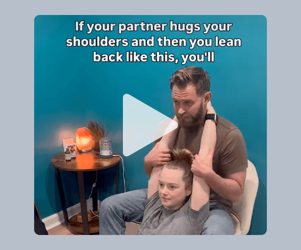 A video still showing a couple demonstrating a stretch, with one person sitting while their partner hugs their shoulders from behind as on-screen text introduces a relationship or wellness tip.