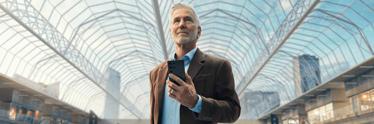 Photo of a silver-haired man in a blazer standing inside a bright, glass-covered atrium, holding his phone and looking thoughtfully ahead. The airy, modern architecture and city buildings in the background give it a calm.