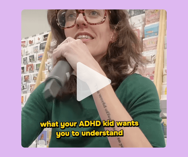 A woman with short brown hair holds a microphone close to her mouth, wearing glasses and a green top, with shelves of books or cards behind her. Yellow text appears across the lower part of the image.