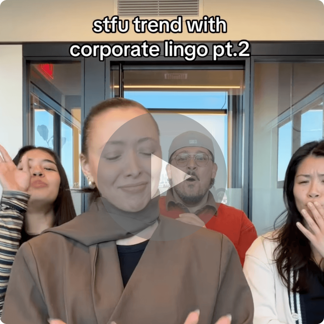 A video still shows four coworkers standing in a glass office space, reacting playfully with expressions and gestures under the text “stfu trend with corporate lingo pt.2.” The tone feels humorous and relatable to office culture.