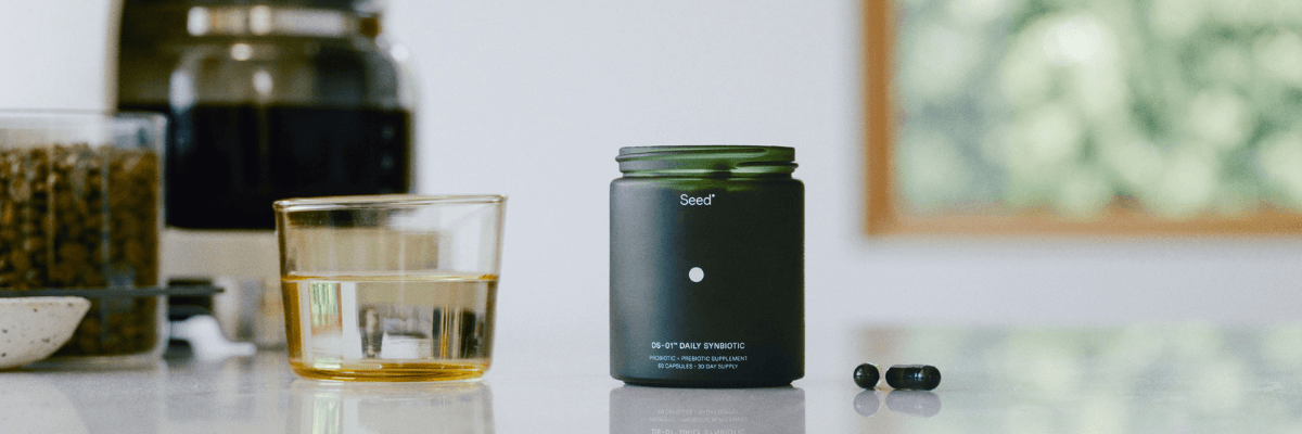 A sleek product shot shows a black jar of Seed daily synbiotic supplements on a kitchen counter beside a glass of water and two capsules. The background is softly blurred with warm, natural light coming through a window.