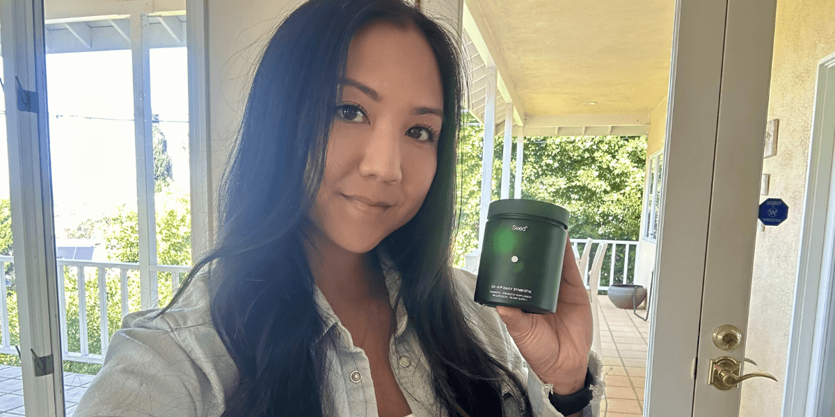A woman stands indoors near a doorway, smiling softly at the camera while holding up a small green container of Seed supplements. Sunlight streams in from the patio behind her, with greenery and a porch visible through the glass door. 
