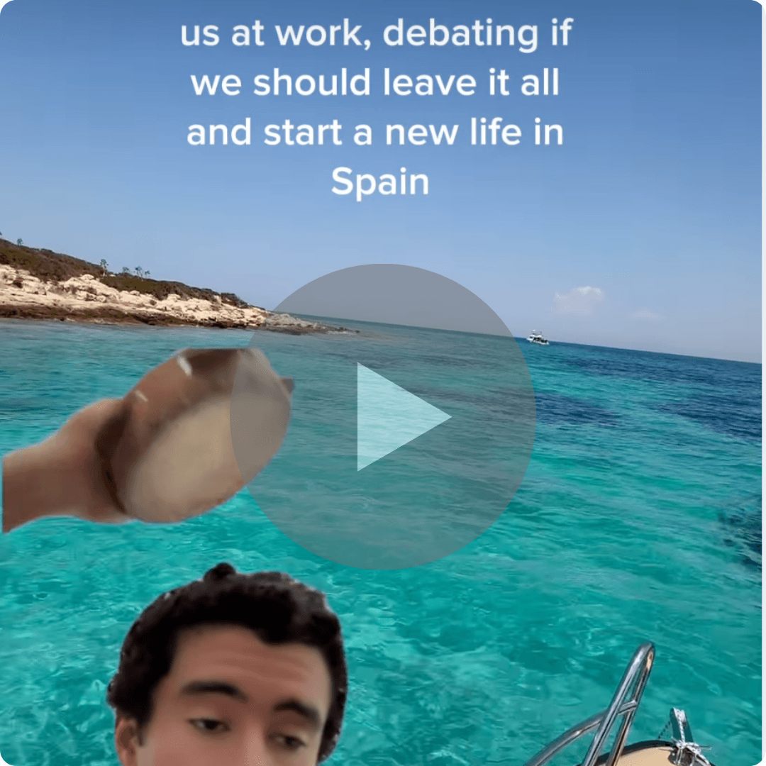 A video thumbnail captures a sunny ocean scene with bright turquoise water and a rocky shoreline in the distance. Overlay text reads, “us at work, debating if we should leave it all and start a new life in Spain,” adding a humorous, escapist tone.