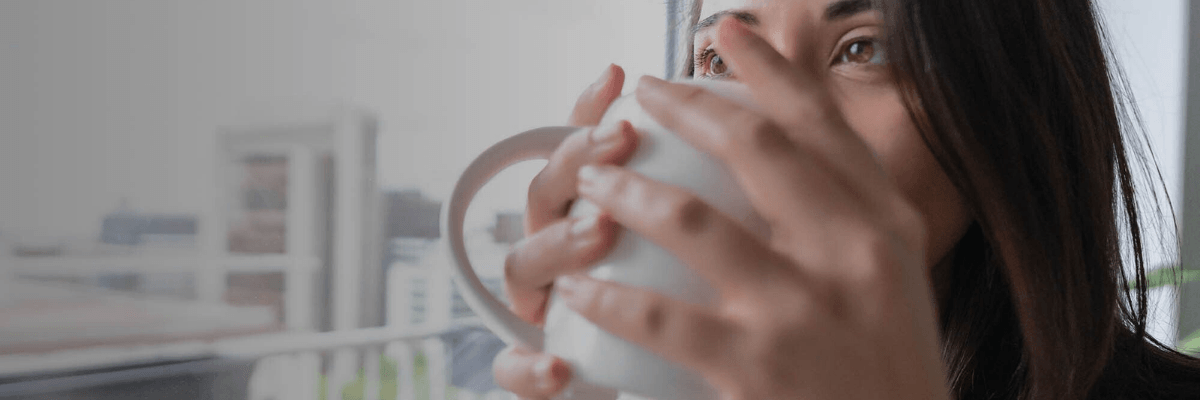 A woman with long dark hair holds a white mug close to her face while looking out a bright window. The background is softly blurred, creating a calm and reflective mood.