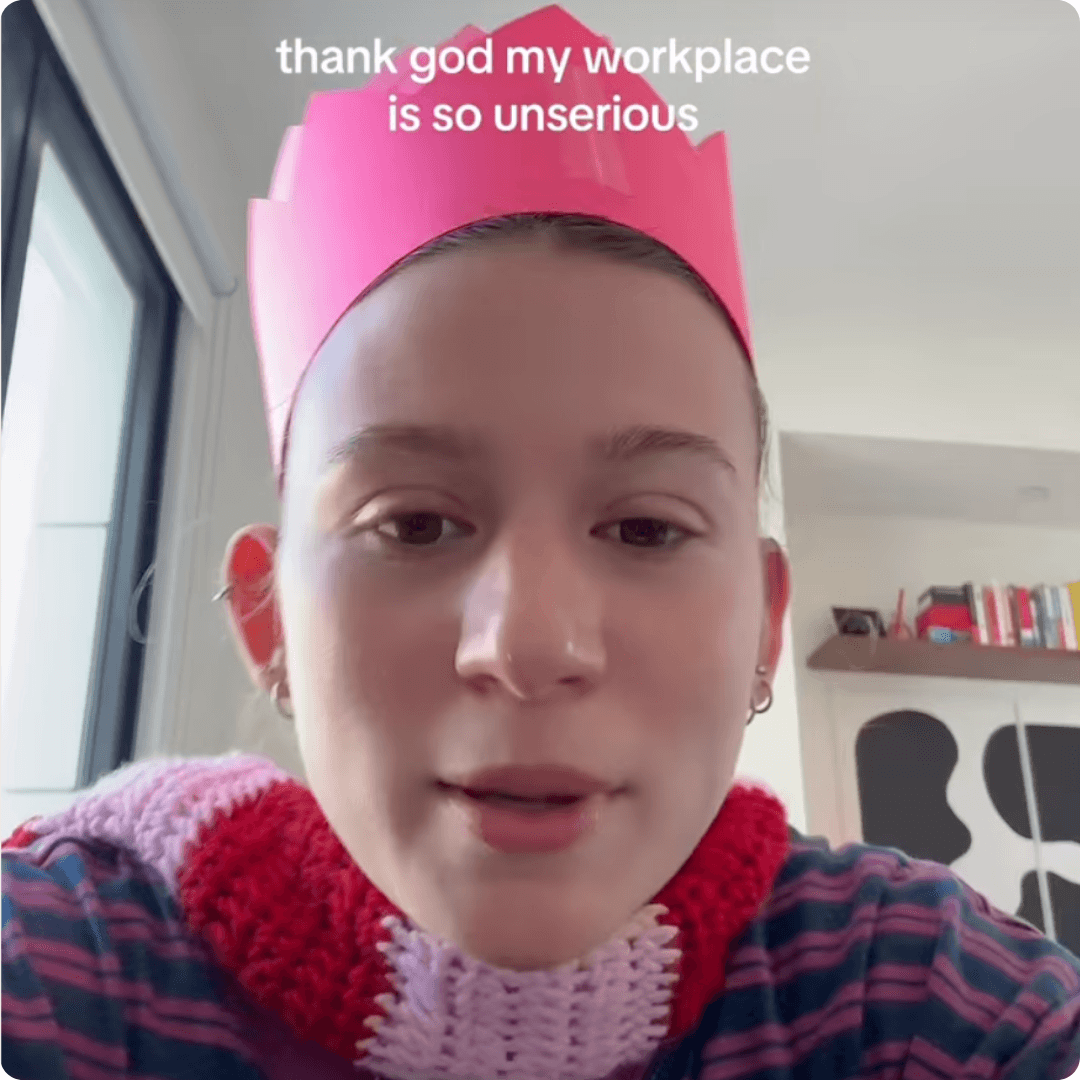 Video screenshot of a person wearing a pink paper crown with text that reads, “thank god my workplace is so unserious.”