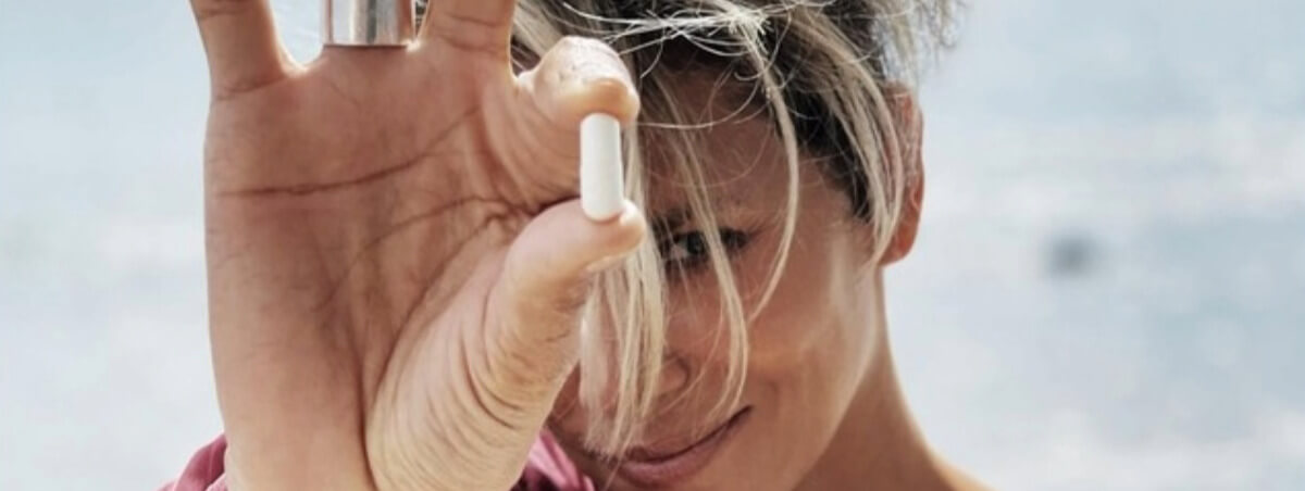 A person holds a white capsule up to the camera while smiling, with windswept hair partially covering their face. The close-up photo has a bright, airy feel and keeps the focus on the supplement in their hand.