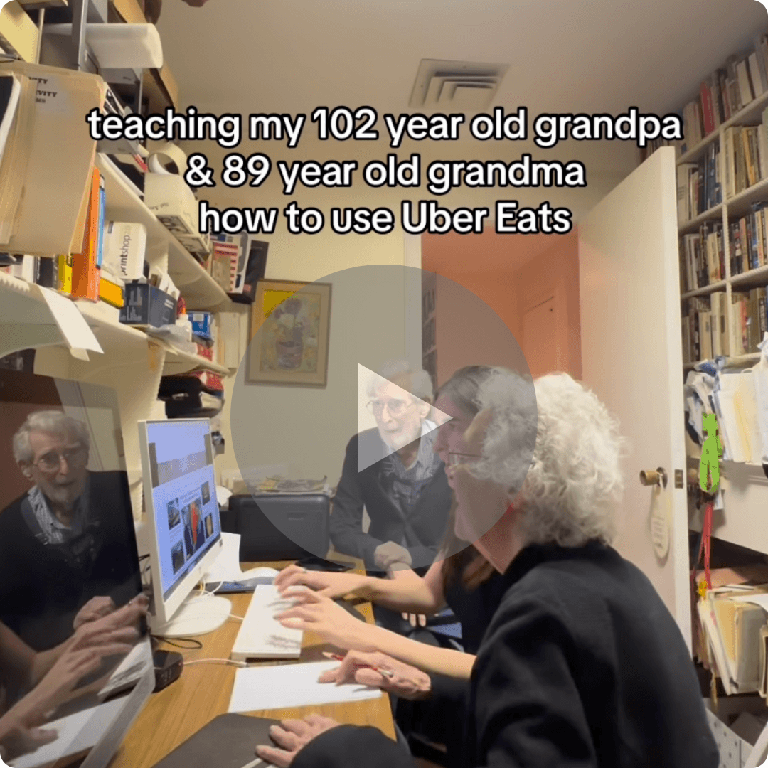 An older couple sits at a computer while a family member helps them navigate a food delivery app. The scene highlights the challenges and humor of learning new technology across generations.