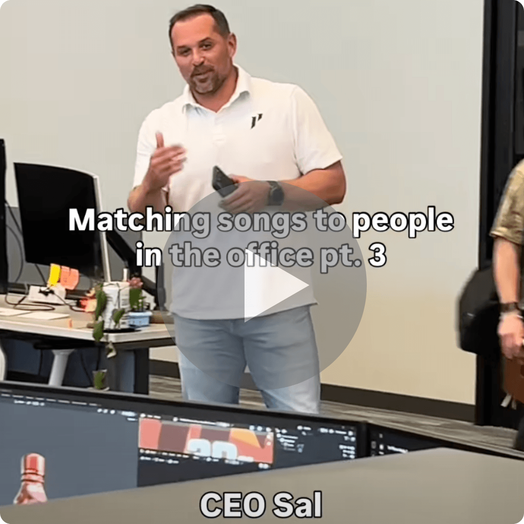 A man stands in an office speaking to coworkers while a caption references matching songs to different people. The video appears to be part of a lighthearted workplace series.