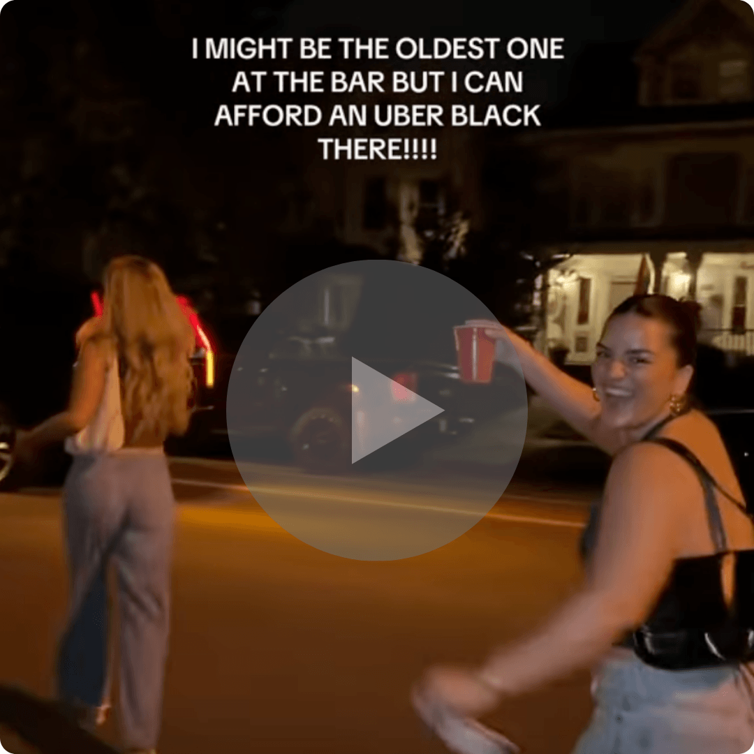 Two women walk across a street at night while smiling and gesturing toward the camera. Houses, parked cars, and streetlights are visible in the background, with overlaid text and a video play icon centered on the image.