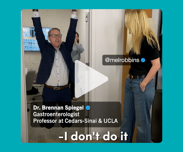 A man in a suit hangs from the top of a doorway with both arms raised while another woman watches and smiles in an office hallway. Text on the video identifies him as a gastroenterologist and professor, with a subtitle at the bottom reading, “I don't do it.”