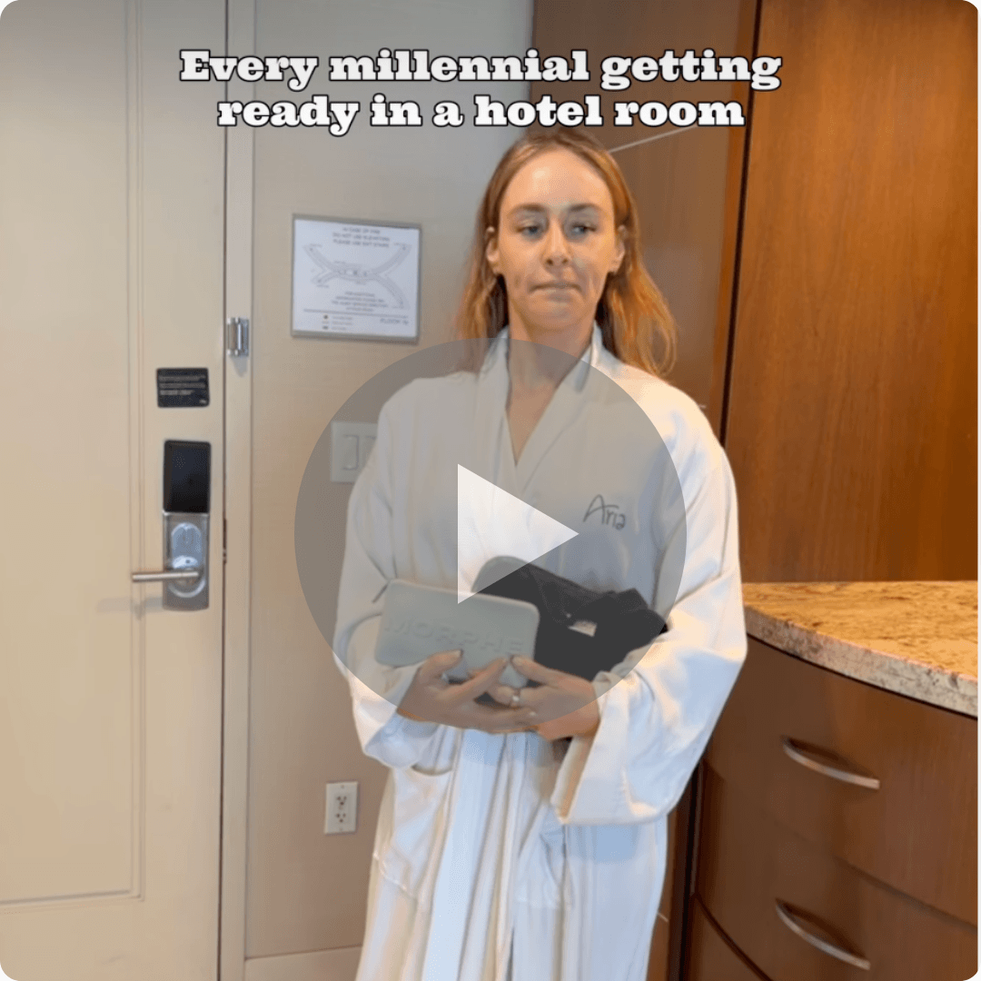 A woman wearing a white hotel robe stands in a hotel room holding several personal items while looking straight ahead. Text at the top reads, "Every millennial getting ready in a hotel room.”