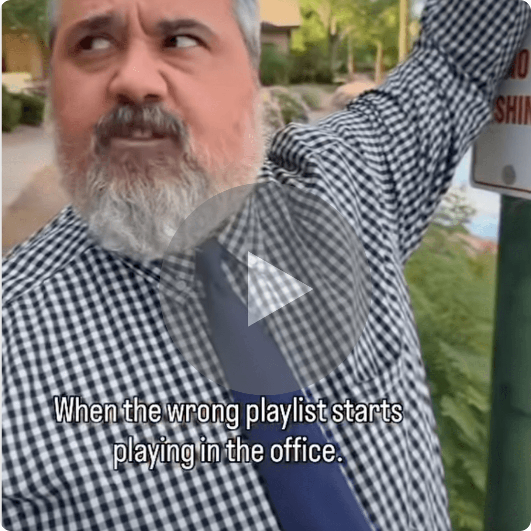 A video thumbnail shows a bearded man in a checkered shirt pointing toward something off-screen outdoors. A caption at the bottom reads, "When the wrong playlist starts playing in the office," with a play button over the center of the image.