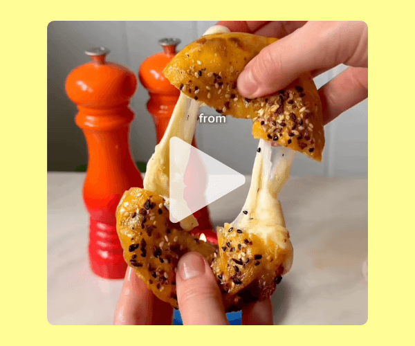A video thumbnail shows hands pulling apart a toasted, cheese-filled bagel with melted cheese stretching between the halves. Two bright orange pepper grinders are blurred in the background on a kitchen counter.