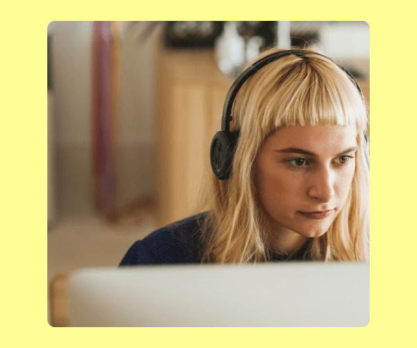 A person with blonde hair and bangs wears over-ear headphones while looking intently at a computer screen. The softly blurred background suggests a home or office workspace.
