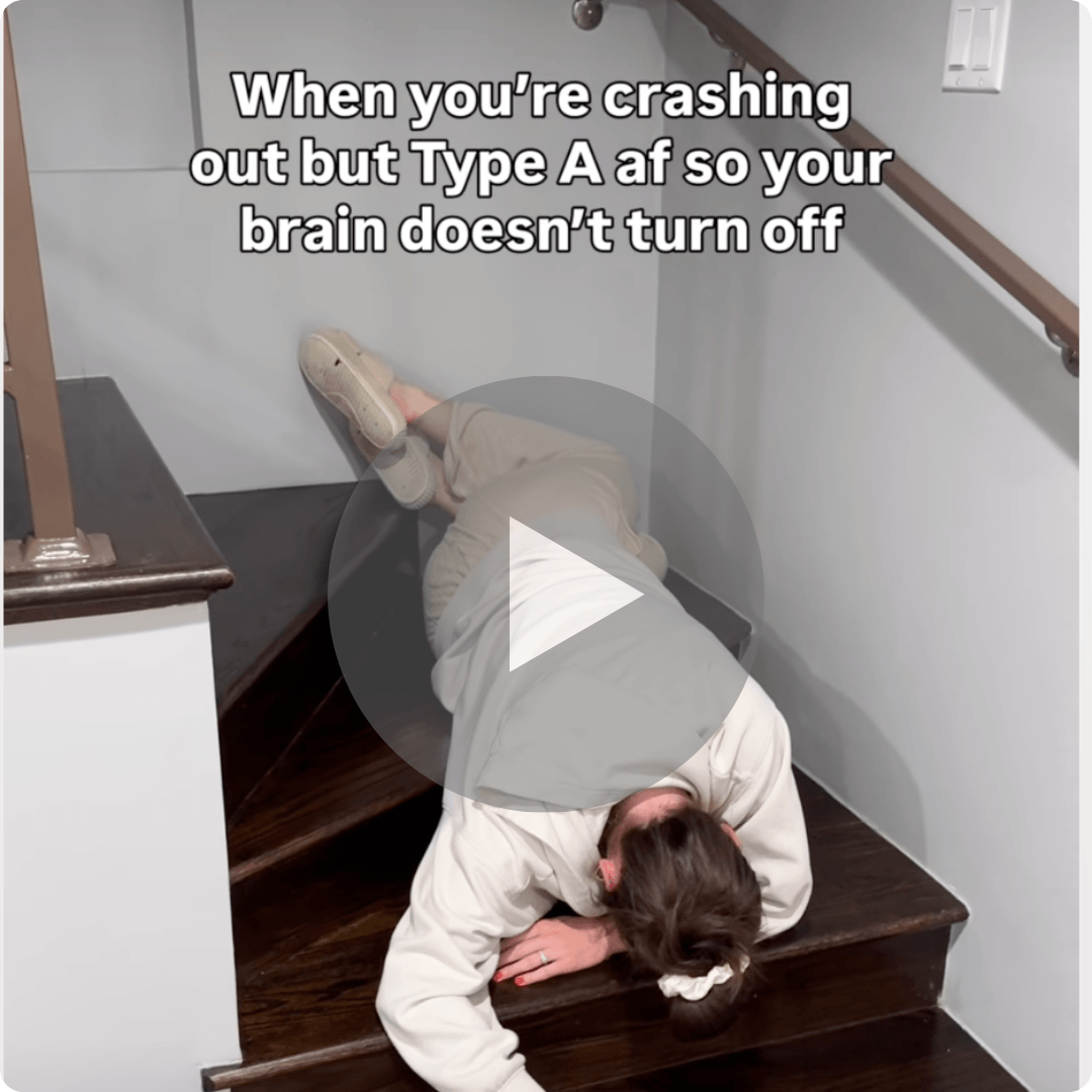 A person in light-colored clothes is sprawled across a dark wooden staircase with their legs resting on the steps. Text at the top reads, “When you’re crashing out but Type A af so your brain doesn’t turn off,” with a large play button over the center.