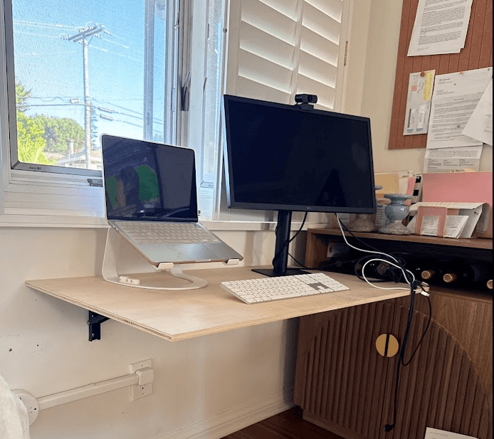 A home workspace features a wall-mounted wooden desk beside a window and dark wood cabinet. A laptop on a raised stand, external monitor, keyboard, webcam, and several cables are arranged across the desk.