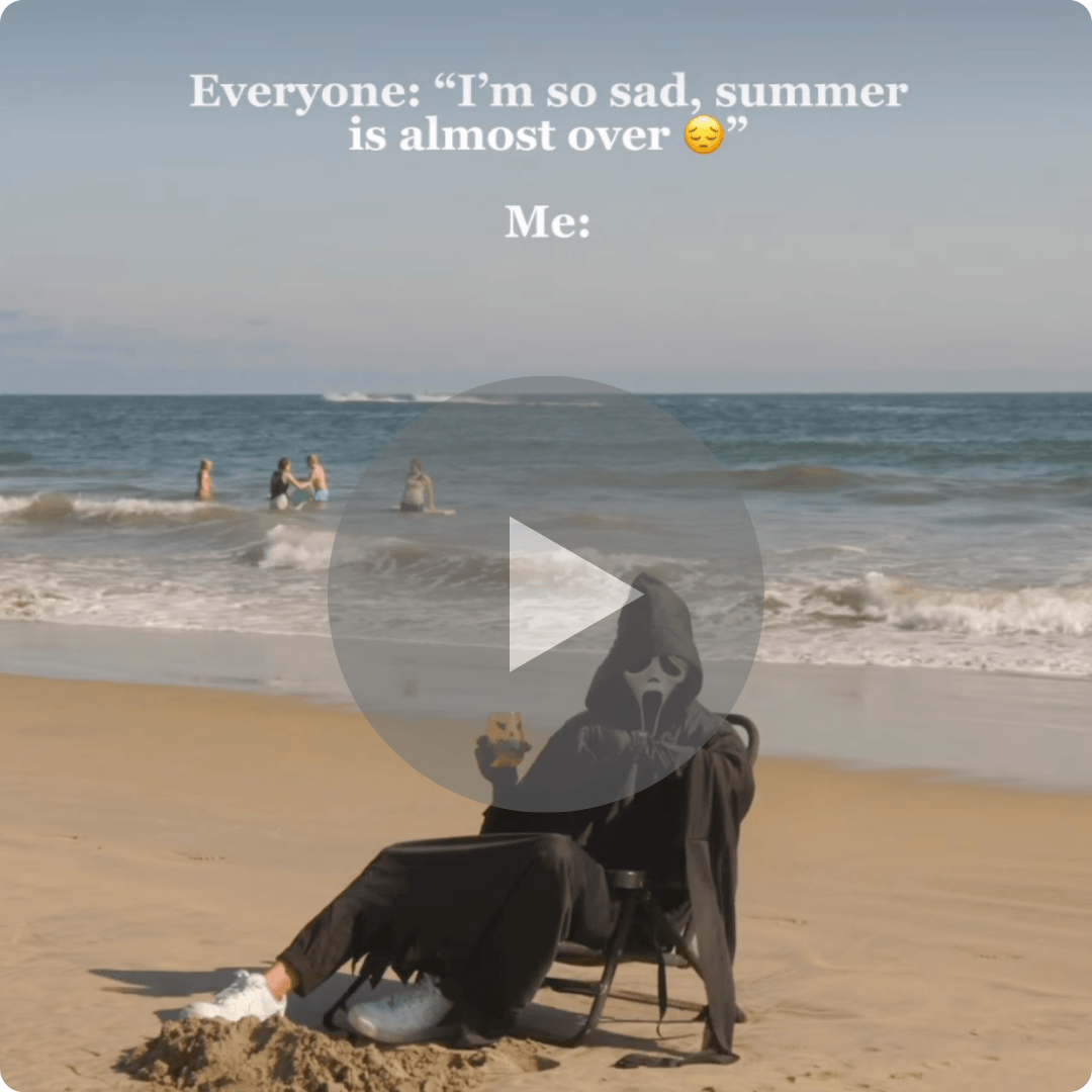 A beach scene shows a person in a black hooded costume sitting in a chair on the sand, with the ocean and several people in the water behind them. White text at the top reads, “Everyone: ‘I’m so sad, summer is almost over’ Me:” and a large play button overlays the center of the image.