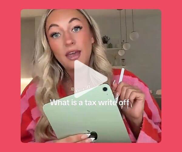 A video-style image with a coral pink background shows a blonde woman holding a light green tablet and stylus indoors. White text reads, “What is a tax write off,” with a translucent play button centered over the image.