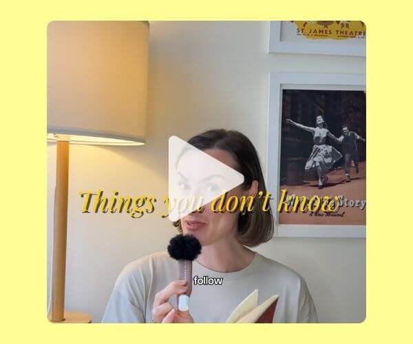 A woman holds a small microphone and a book while speaking to the camera. Framed photographs and a floor lamp are visible behind her, with text overlaid across the image.