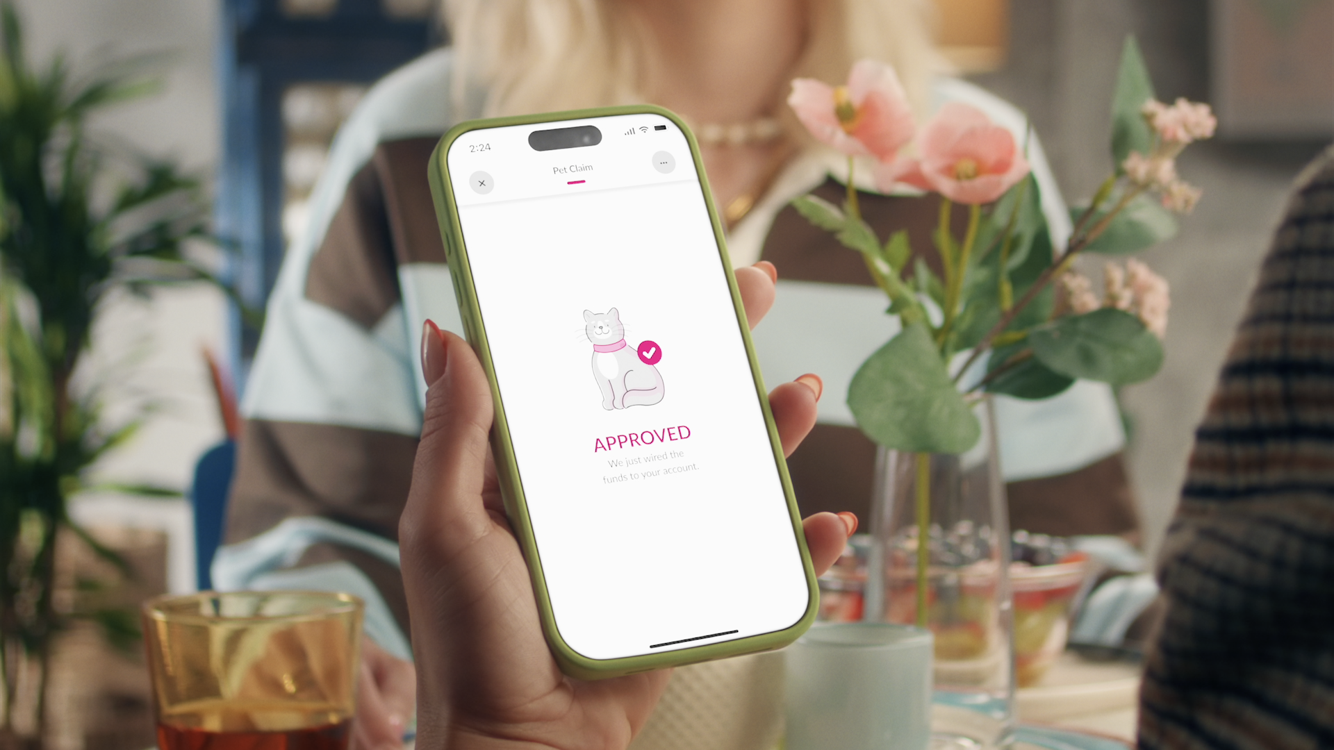 A hand holds a smartphone displaying a “Pet Claim” screen with an illustrated cat and the word “APPROVED.” A person, flowers, drinking glasses, and a table are visible in the background.
