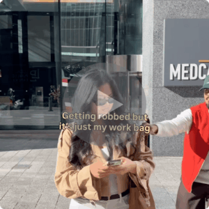 A woman in sunglasses looks at her phone on a sidewalk while another person reaches toward her bag. Overlaid text reads, βGetting robbed but itβs just my work bag,β with a large play button in the center.