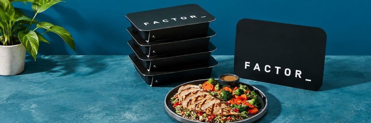 The image shows several black rectangular meal containers stacked beside a plated meal with sliced chicken, vegetables, grains, and a small container of sauce. A large black sign with the word “FACTOR_” stands behind the meal, with a green potted plant on the left.
