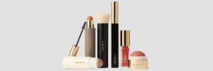 Merit makeup products, including brushes, lip color, and a pink cream blush, are arranged against a light gray background. Their packaging is black, cream, and gold.