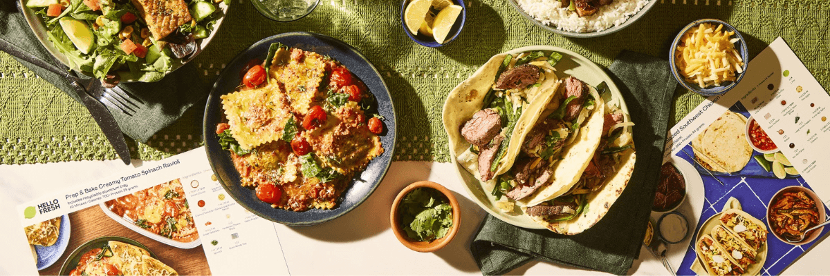 A top-down food spread features colorful dishes including ravioli, tacos, rice, salad, and various side dishes arranged across a textured green tablecloth. Recipe cards and meal-kit packaging are placed around the plates.