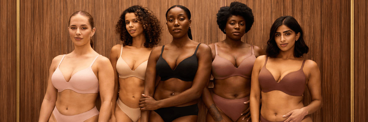 Five women stand side by side against a brown wood-paneled wall. They wear matching bra and underwear sets in shades of pink, beige, black, and brown.