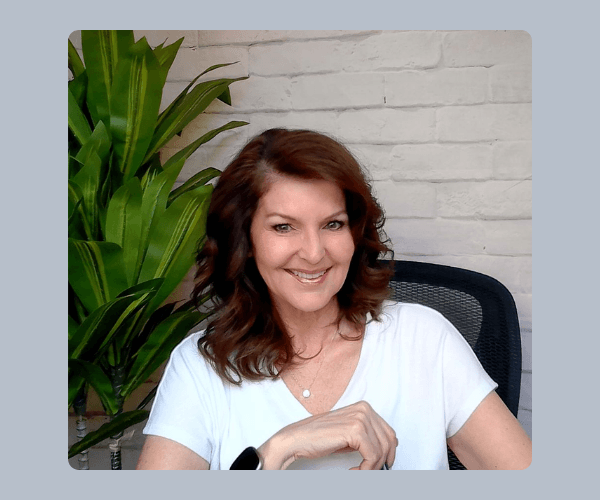 A smiling woman with shoulder-length brown hair wears a white shirt while sitting in a black chair. A green plant and white brick wall are visible behind her.