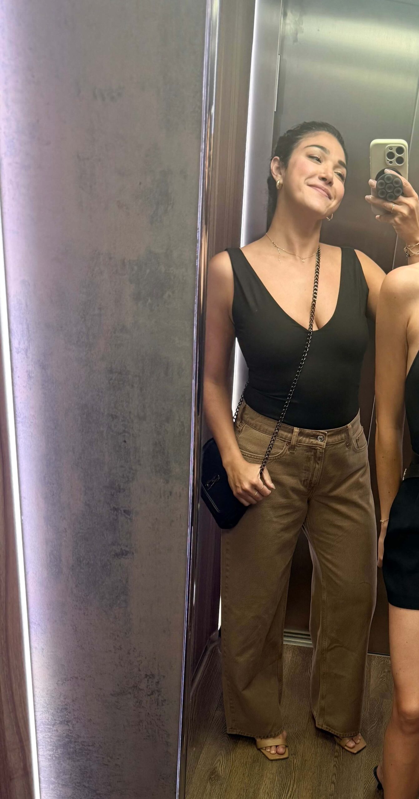 A woman takes a mirror selfie while wearing a black sleeveless V-neck top and brown wide-leg pants. She carries a small black crossbody bag and wears gold jewelry and beige open-toe shoes.