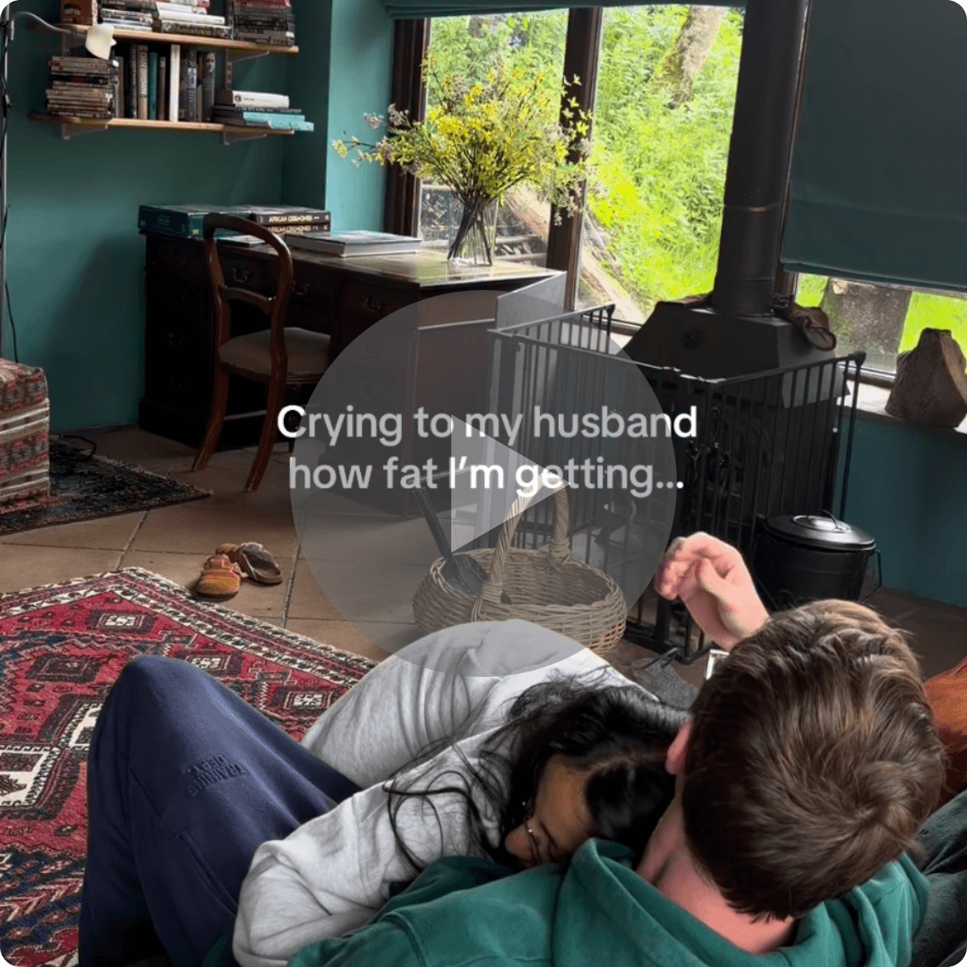 A person rests against another person in a room with teal walls, a desk, bookshelves, large windows, and a black stove. White text over the video reads, “Crying to my husband how fat I’m getting...” with a play button in the center.