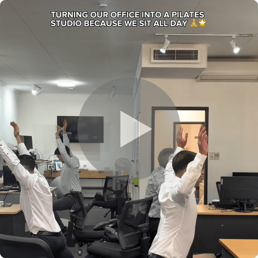 A video thumbnail shows several office workers seated in rolling chairs with their arms raised inside an office with desks and computer monitors. White text at the top reads “TURNING OUR OFFICE INTO A PILATES STUDIO BECAUSE WE SIT ALL DAY,” followed by two emojis.